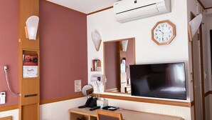 Ein Hotelzimmer mit Schreibtisch, Stuhl, Fernseher, Klimaanlage, Uhr und einem Bücherregal mit Büchern.