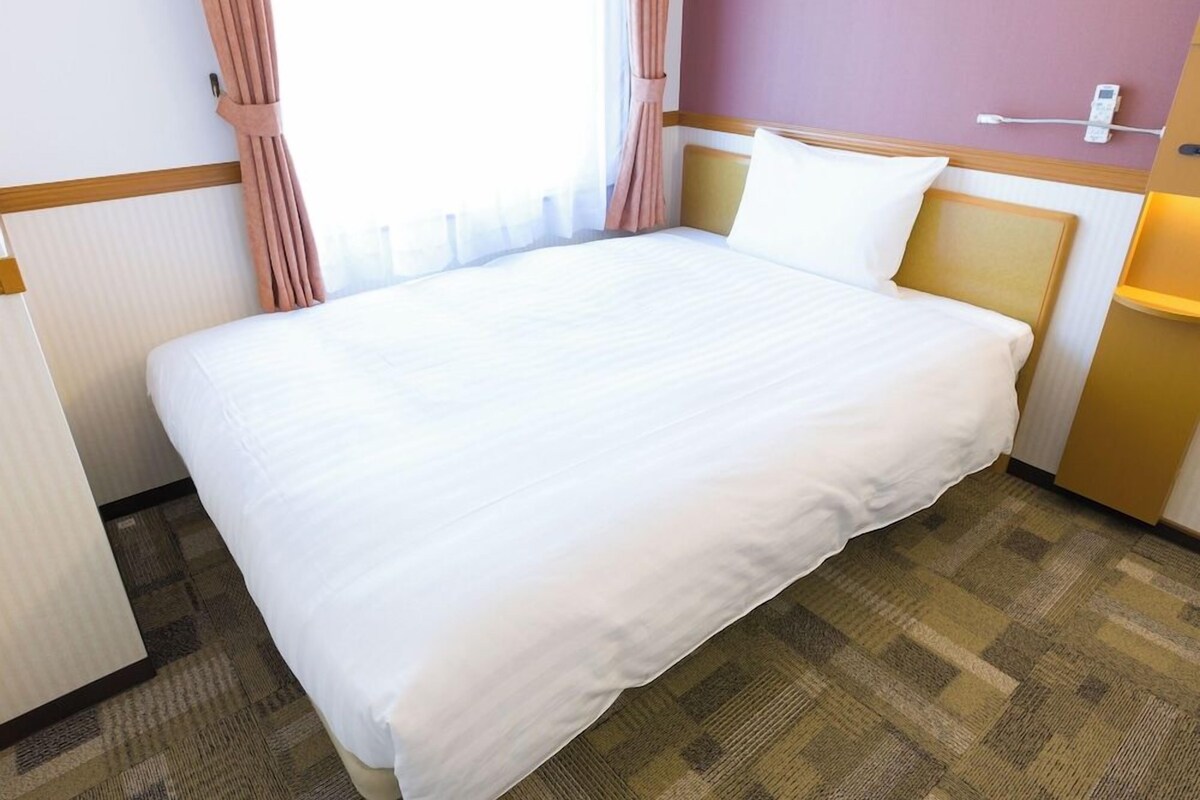 A hotel room with a large bed, a bedside table, a lamp, and a window with curtains.