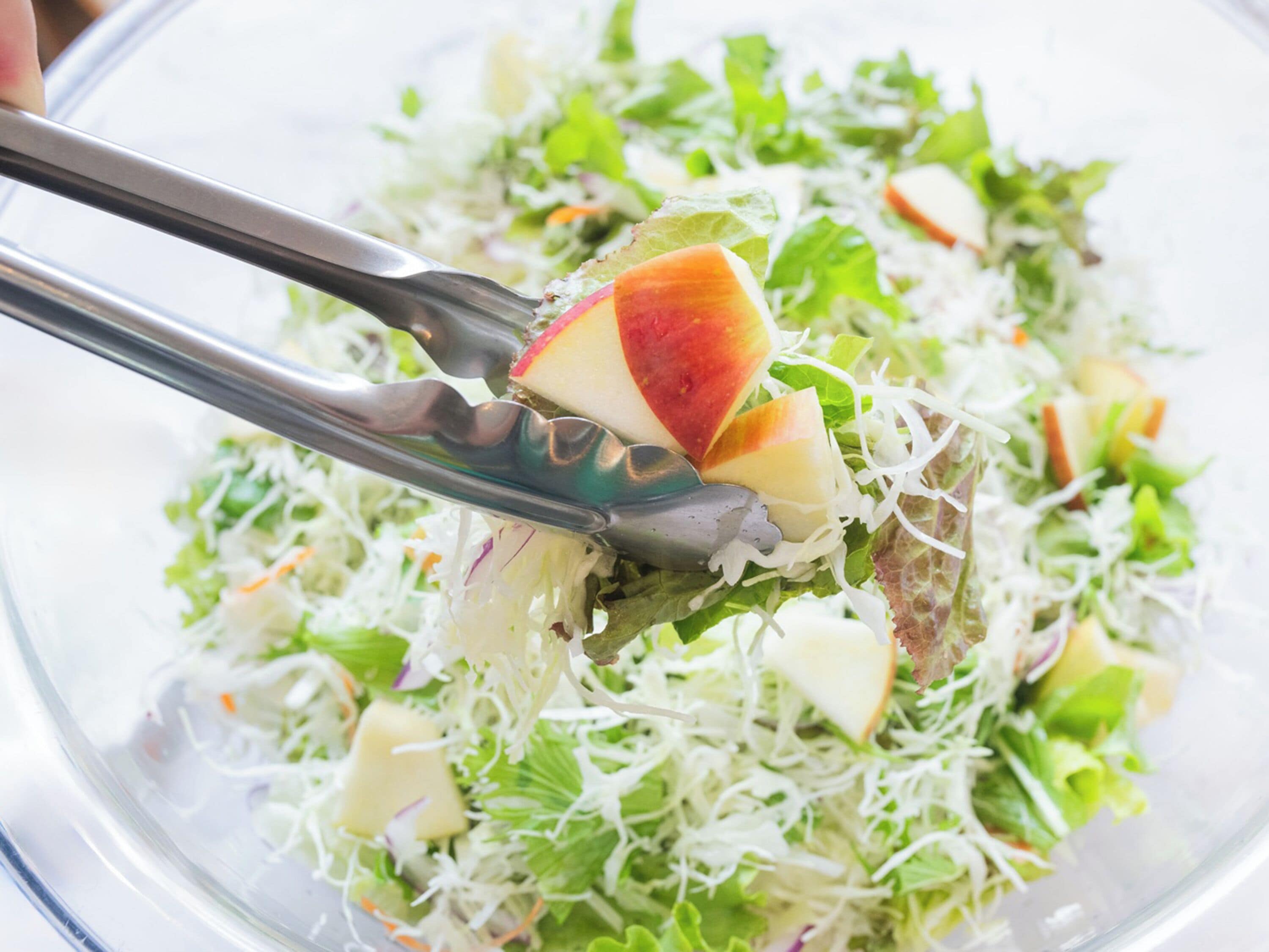 Une salade composée de divers légumes et de morceaux de fruits, présentée dans un saladier et servie à l’aide de pinces.