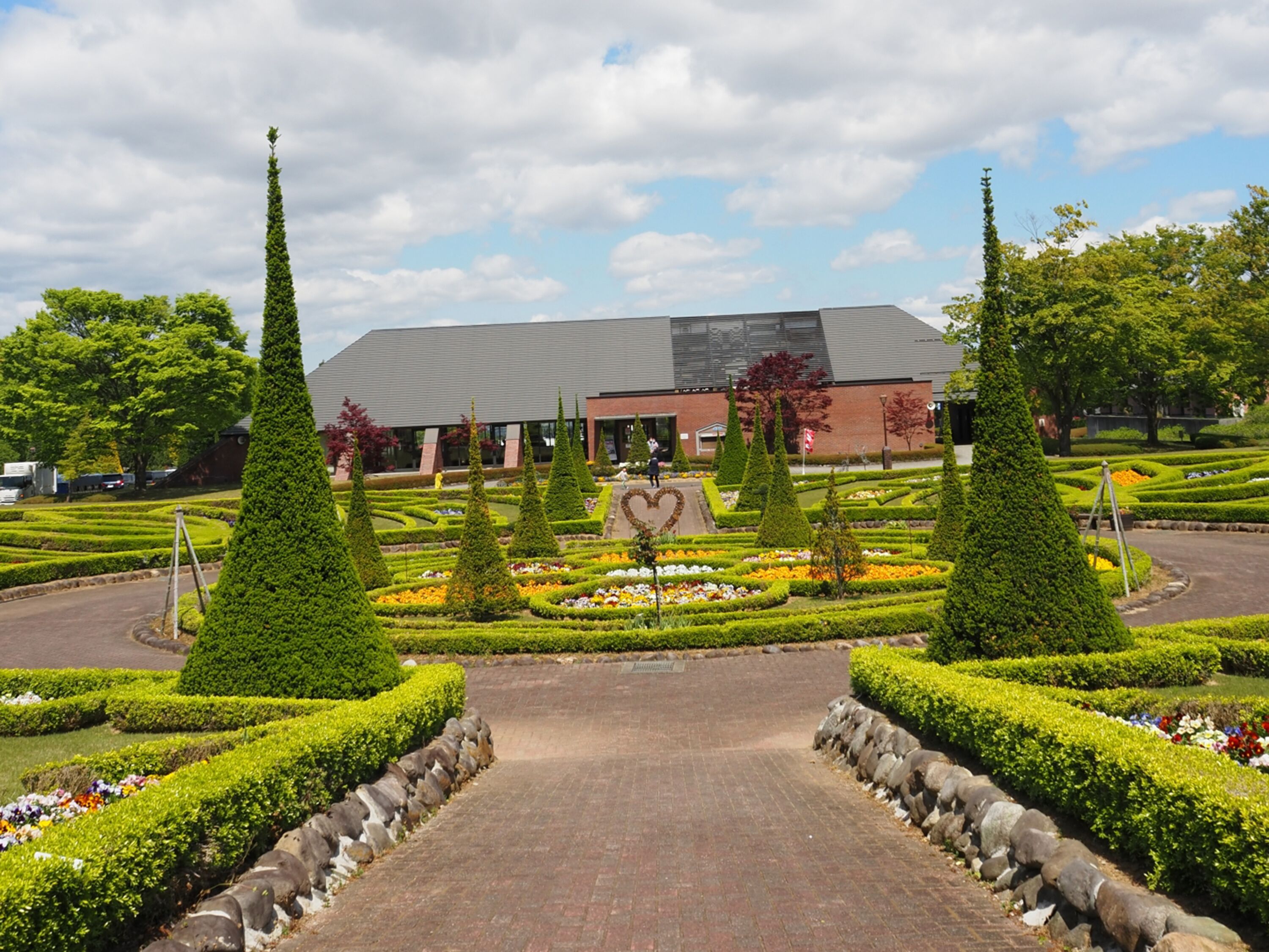 Un bâtiment avec un jardin à la française devant, comprenant des haies soigneusement taillées et un parterre floral en forme de cœur.