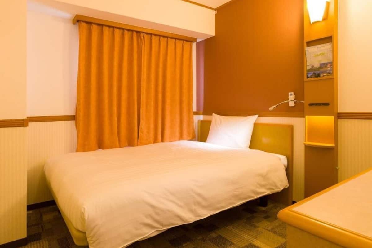 Ein Hotelzimmer mit Bett, kleinem Tisch, Wandlampe und einem Fenster mit Vorhängen.