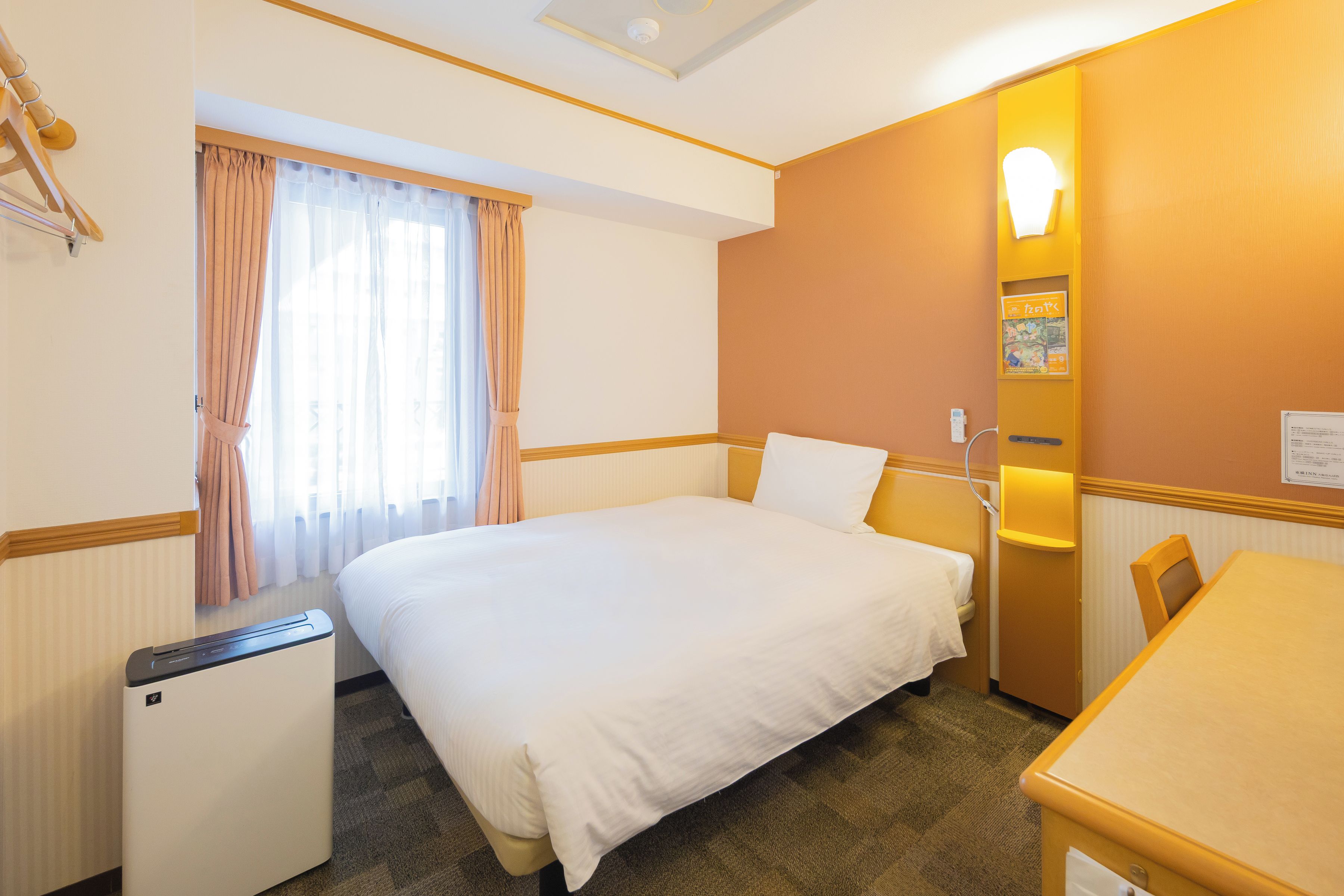 Een hotelkamer met een bed, een bureau, een stoel, een tafeltje, een radiator, een raam met gordijnen en een wandlamp.