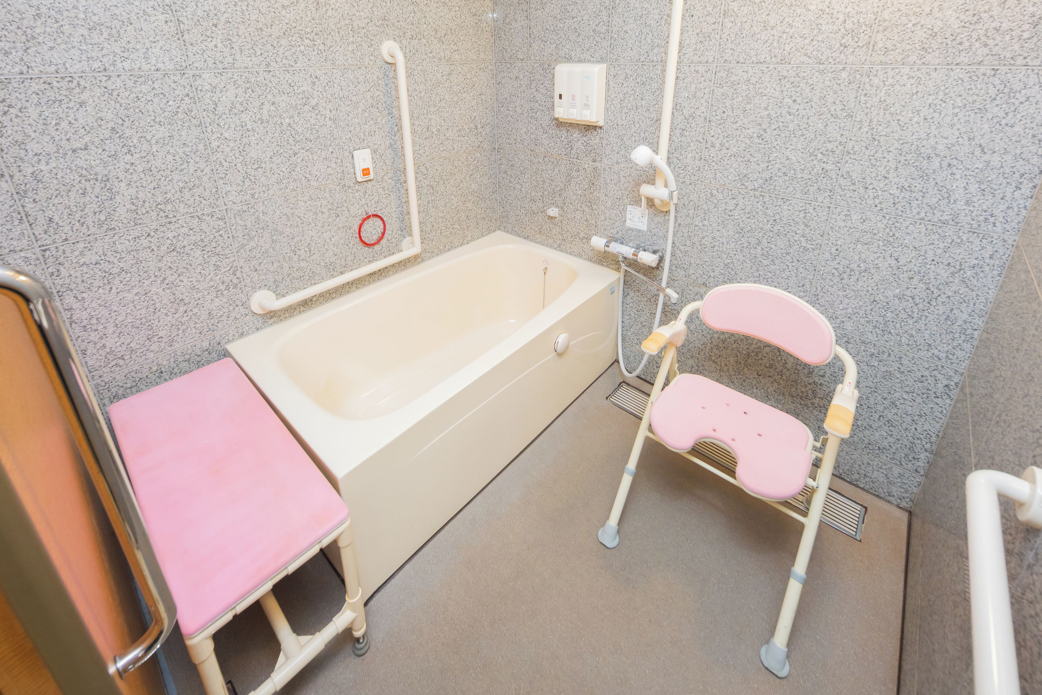 A bathroom with a bathtub, a pink stool, a handrail, and a shower chair.