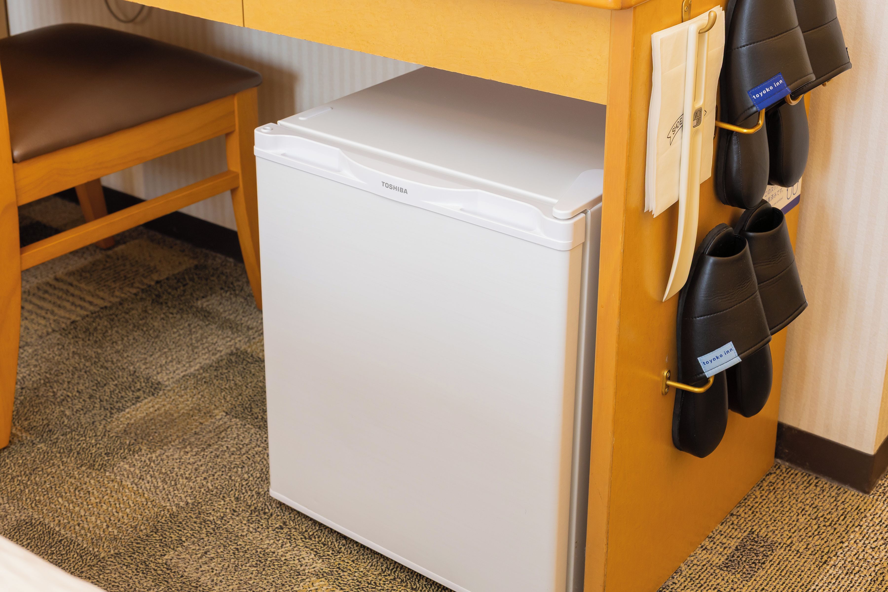 Un refrigerador blanco compacto en una habitación de hotel, con una silla de madera y un zapatero con zapatos negros.