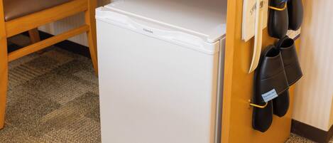 Un refrigerador blanco compacto en una habitación de hotel, con una silla de madera y un zapatero con zapatos negros.