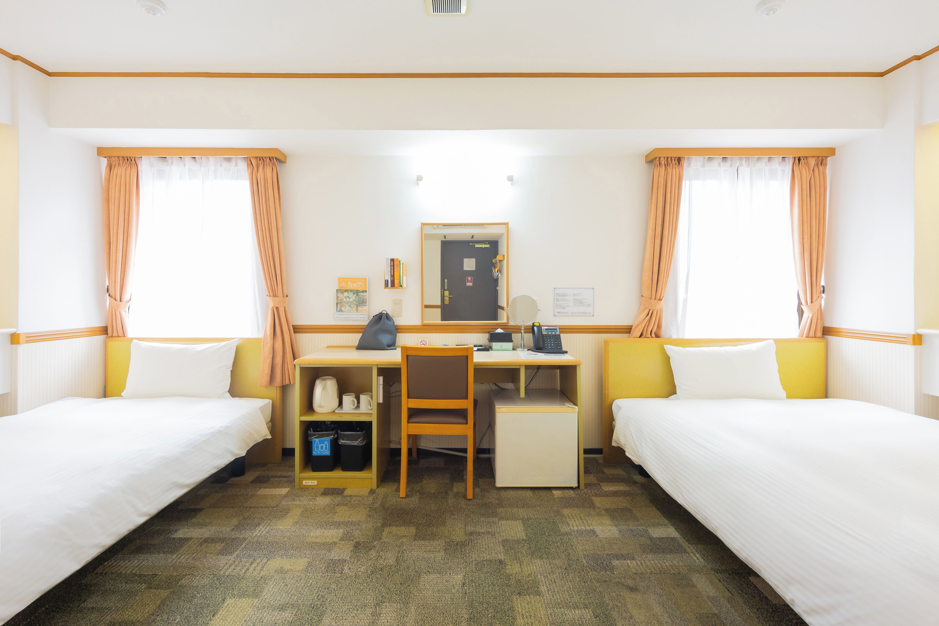 A hotel room with two single beds, a desk with a chair, a telephone, and a small shelf with books.