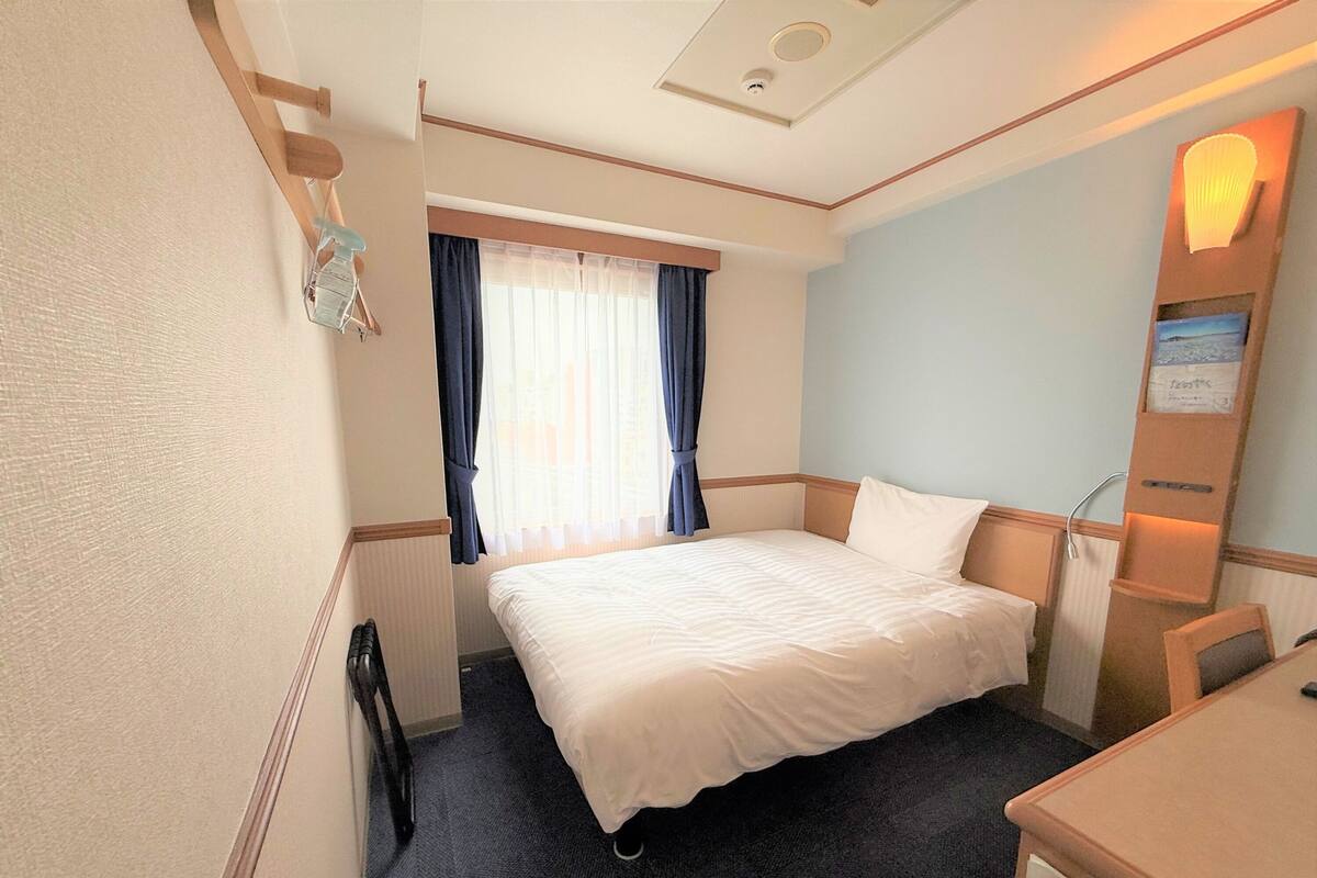 A hotel room with a bed, a desk, a chair, a nightstand, a lamp, a window with curtains, and a wall-mounted shelf with a picture.