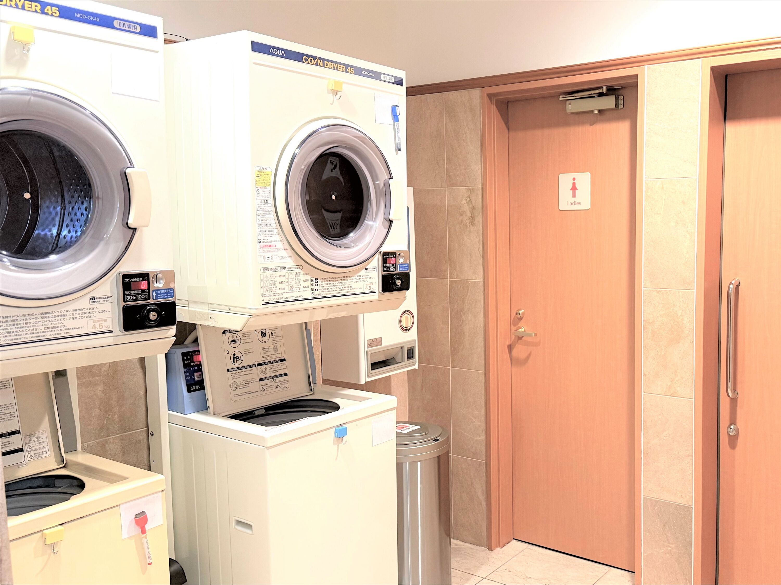 A laundry area with two front-loading washing machines and a trash bin.