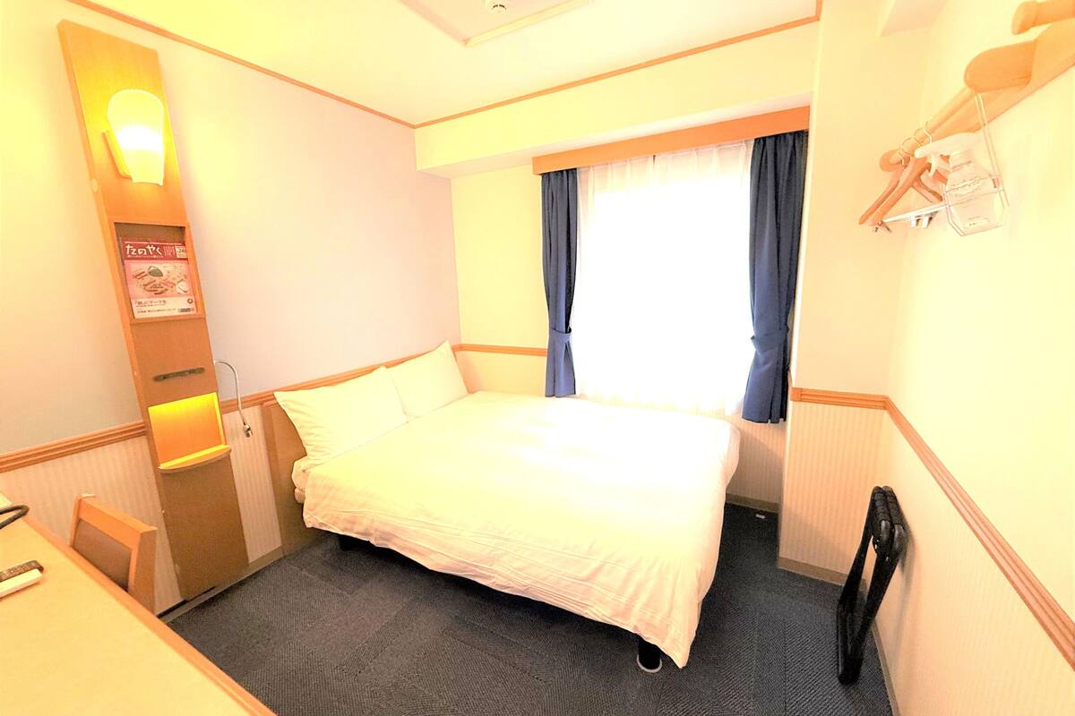 A hotel room with a bed, a desk, a chair, a window with curtains, and a wall-mounted TV.