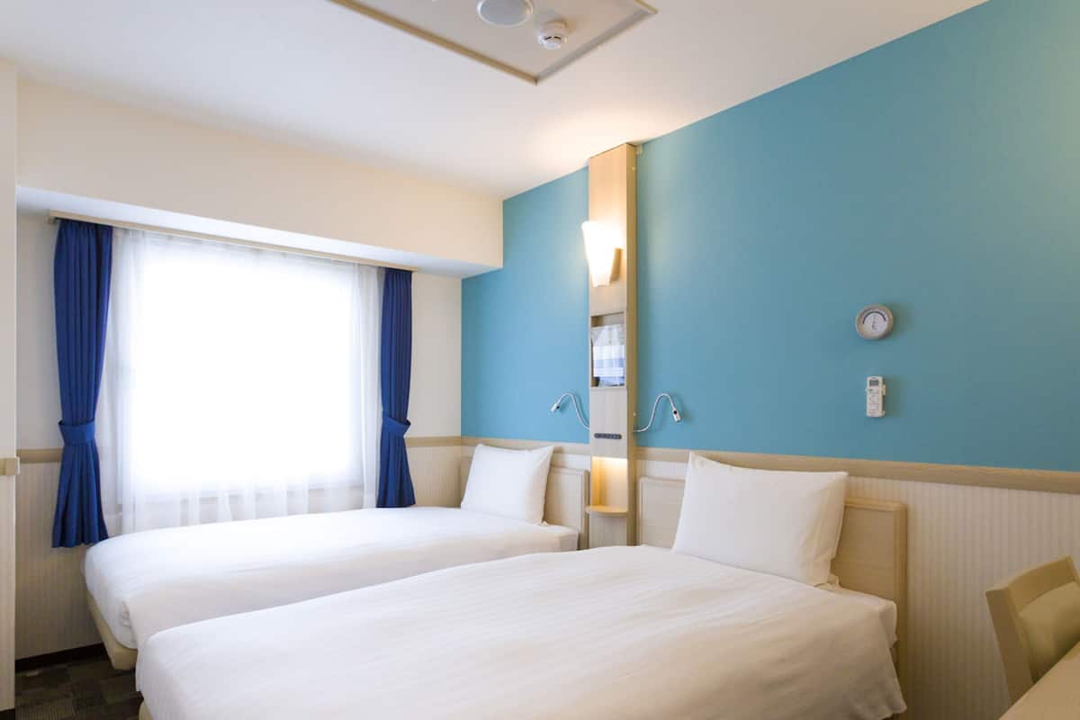 A hotel room with two beds, a window with curtains, a wall clock, and a thermostat.