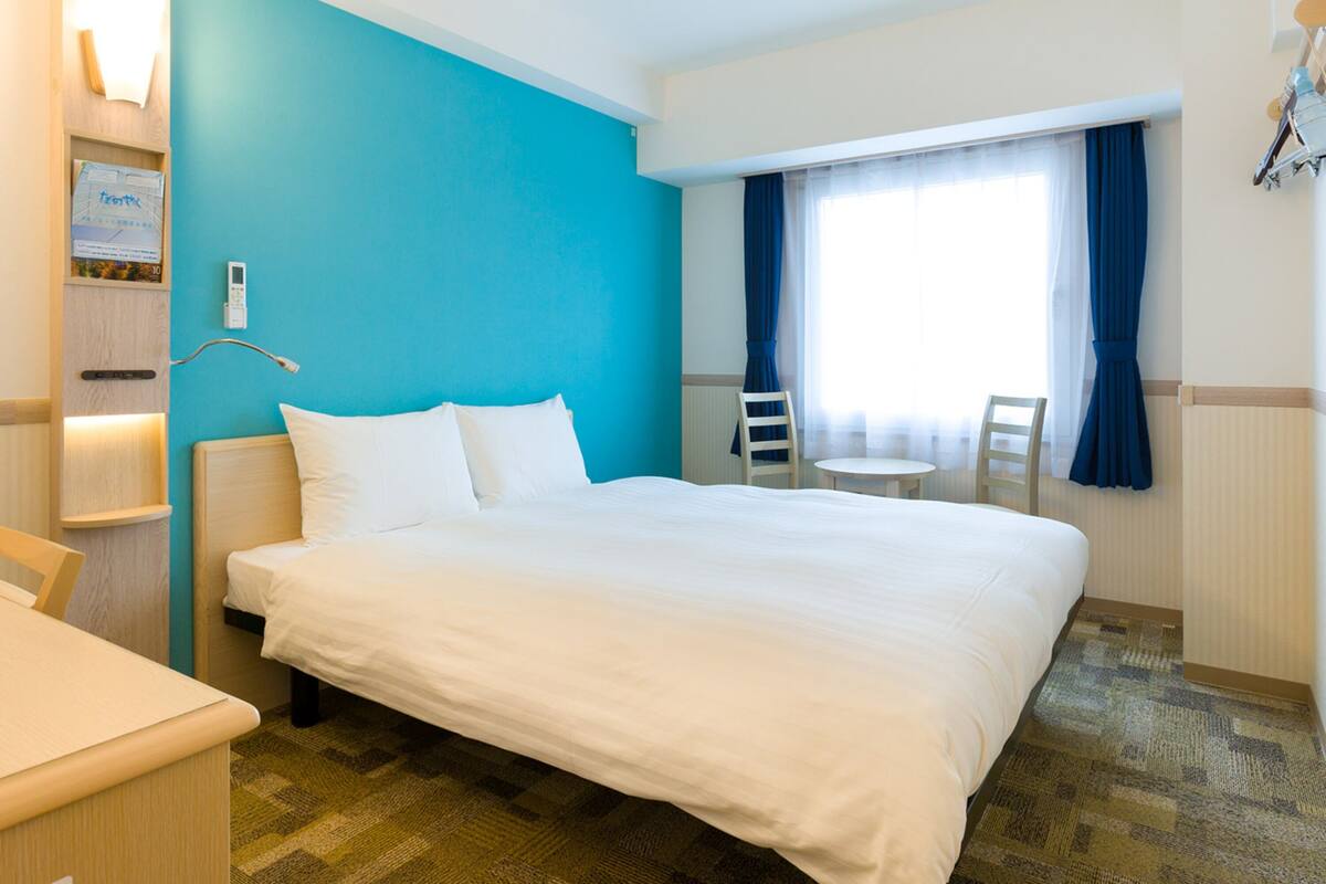 A hotel room with a large bed, a bedside table, a chair, a small table, and a window with curtains.