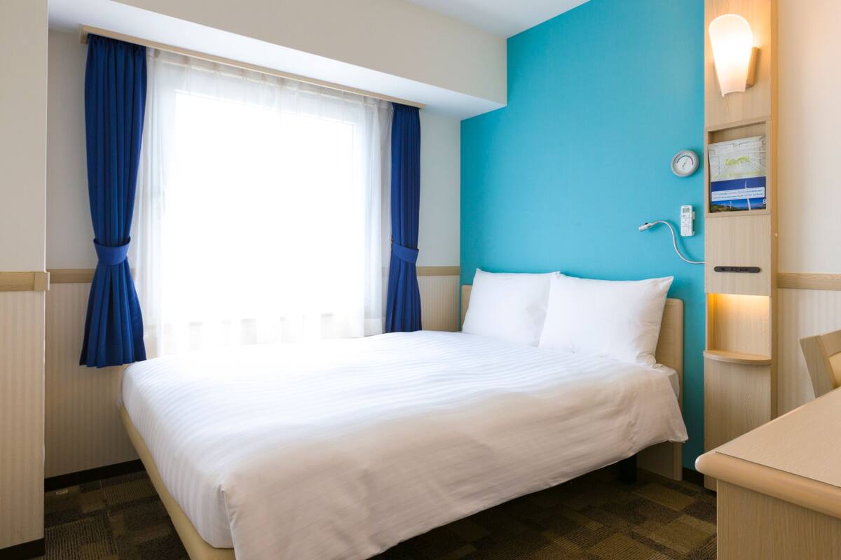 A hotel room with a bed, a desk, a wall-mounted clock, and a window with curtains.