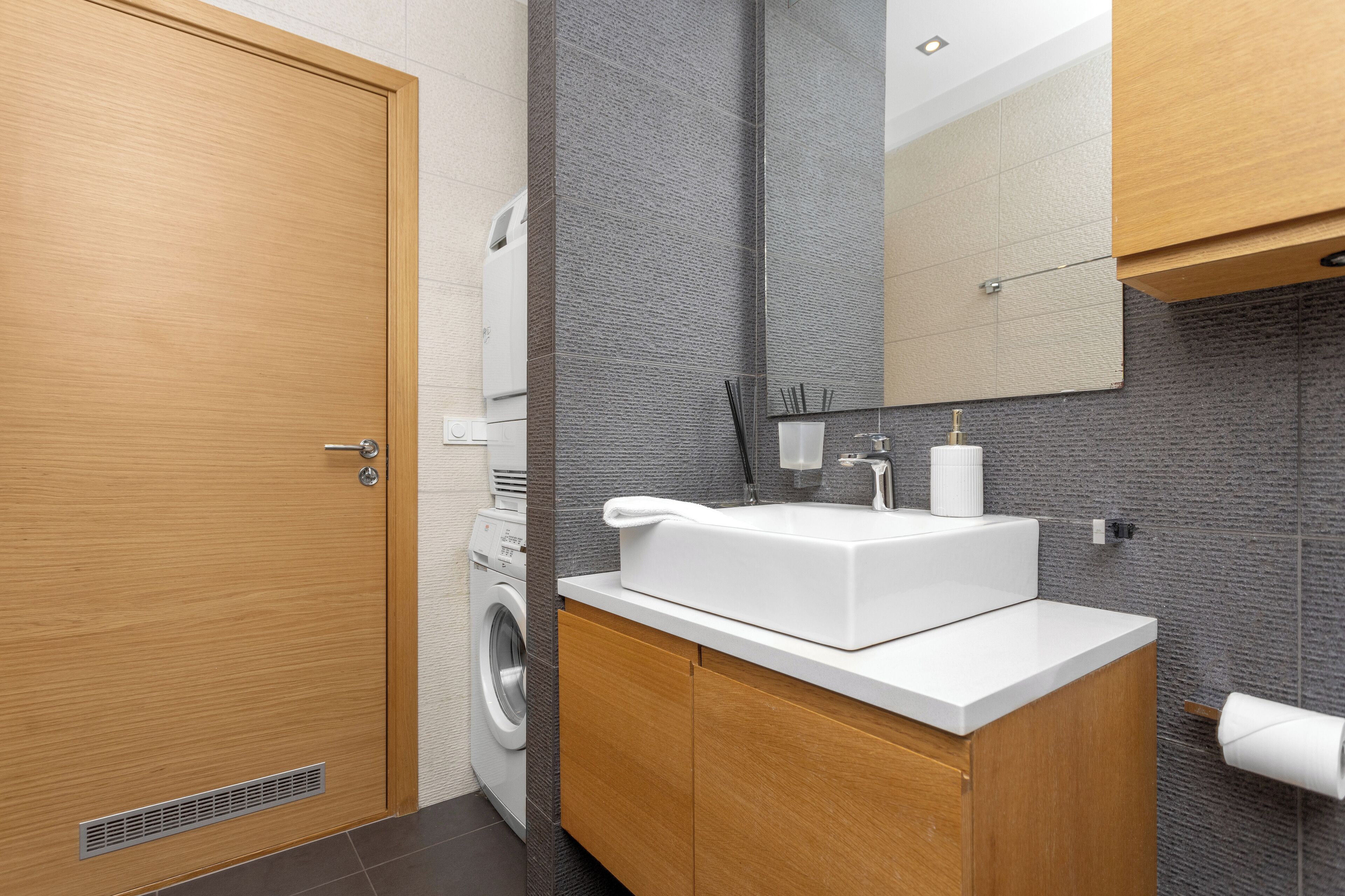 Une salle de bain moderne avec une porte en bois, un lave-linge, un lavabo blanc et un grand miroir.