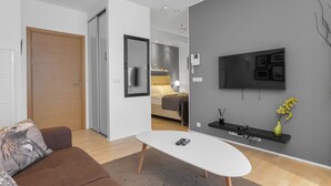 A modern living room with a sofa, a coffee table, a TV mounted on the wall, and a hallway leading to a bedroom.