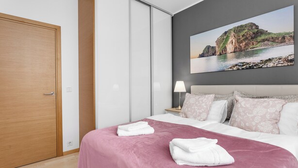 A neatly made bed with a pink bedspread and white pillows, a wooden door, a nightstand with a lamp, and a large painting of a coastal landscape on the wall.