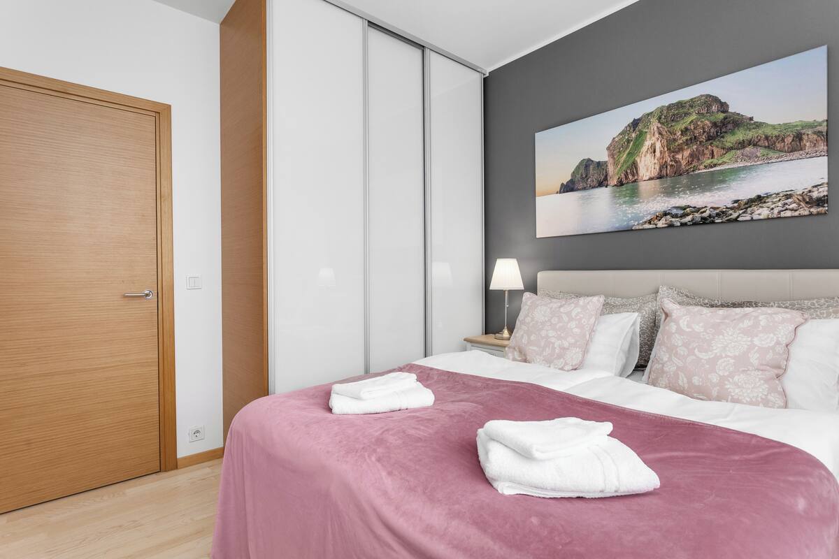 Ein ordentlich bezogenes Bett mit einer rosa Bettdecke und weißen Kissen, eine Holztür, ein Nachttisch mit Lampe und ein großes Gemälde einer Küstenlandschaft an der Wand.