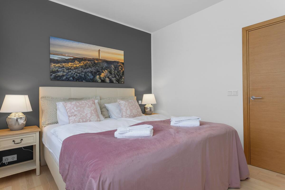 Ein ordentlich gemachtes Bett mit einer rosa Decke, zwei Nachttischlampen und einem Gemälde mit einer Küstenlandschaft an der Wand.