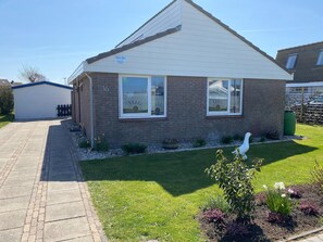 A single-story house with a brick facade, two windows with white frames, a small garden with grass and flowers, and a white bird statue.