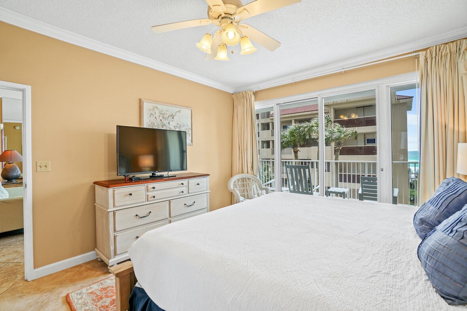A hotel room with a large bed, a television on a dresser, a balcony with a view of the ocean, and a ceiling fan.