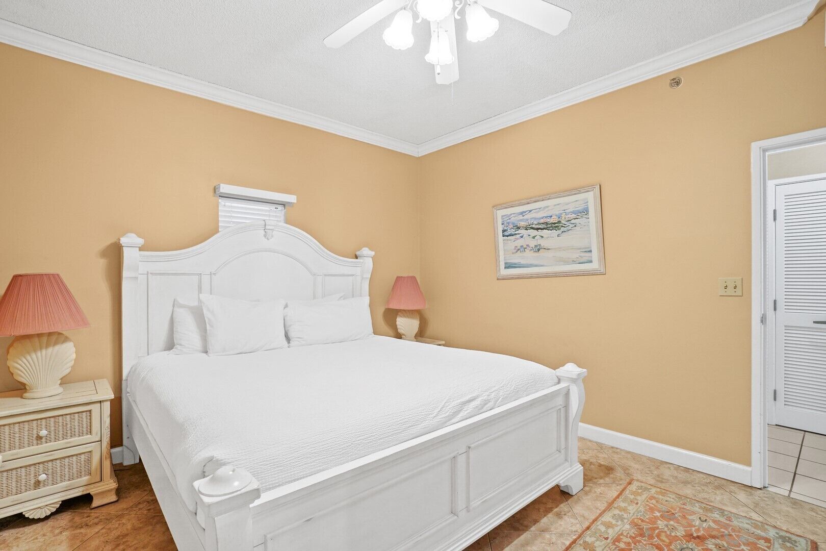 Un dormitorio con una cama blanca, dos lámparas de noche, un ventilador de techo blanco, una pared color beige y un cuadro enmarcado.