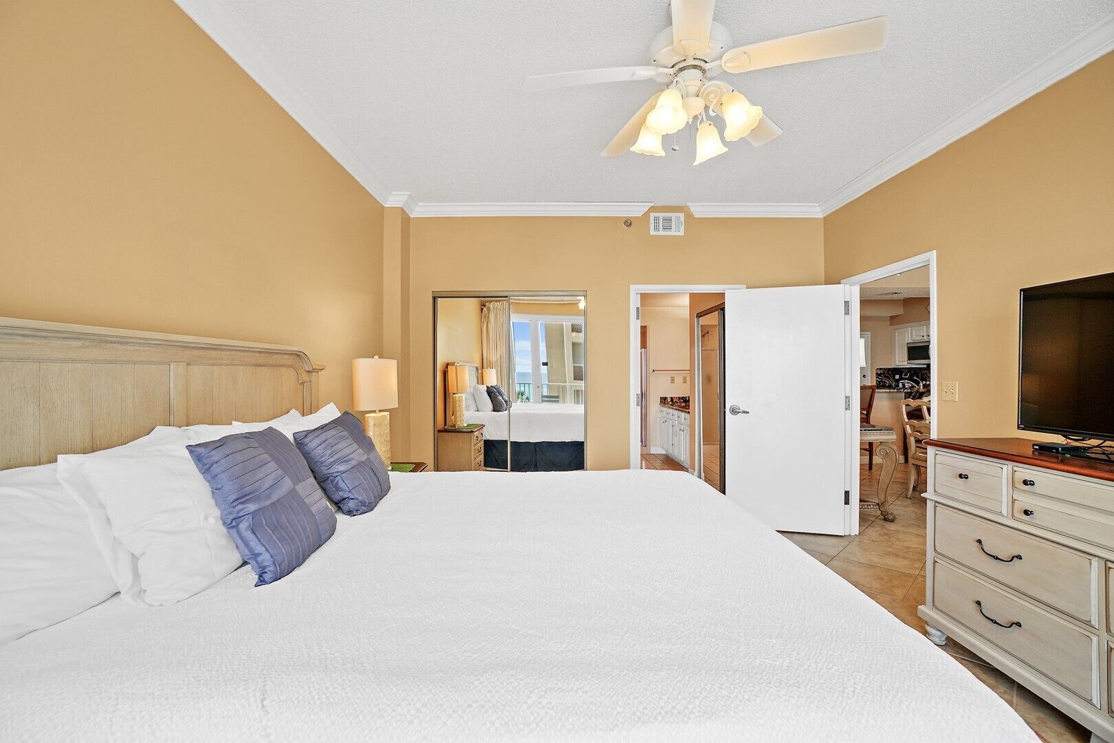 A hotel room with a large bed, a ceiling fan, a television, and a view of the bathroom and kitchen through an open door.