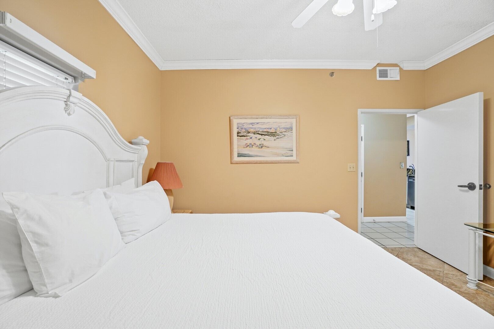 A bedroom with a white bed, a painting on the wall, and a door leading to another room.