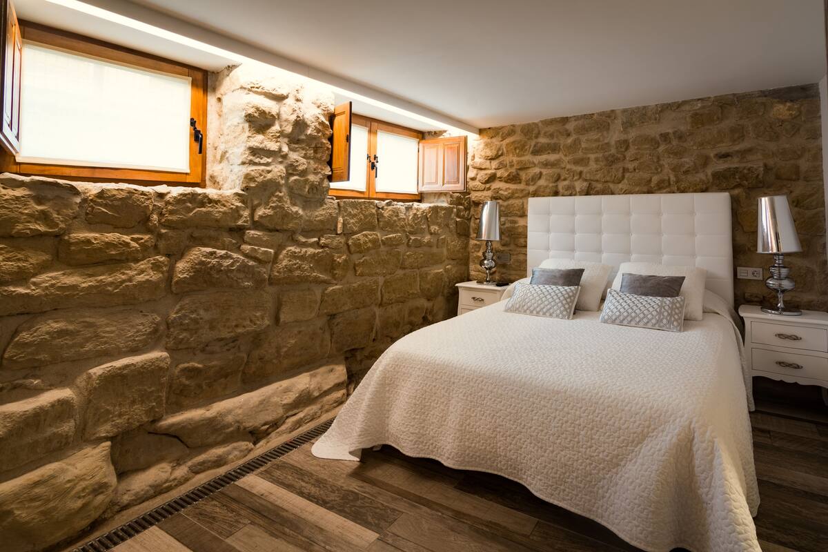 Quarto com parede de pedra, cama com cabeceira branca e piso de madeira.