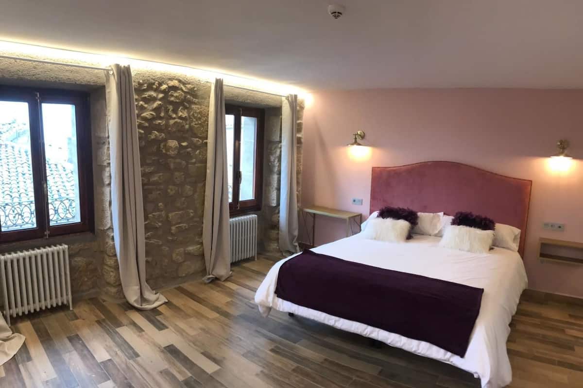 Quarto com parede de pedra, cama com roupa de cama branca e roxa, duas luminárias de cabeceira e um radiador.