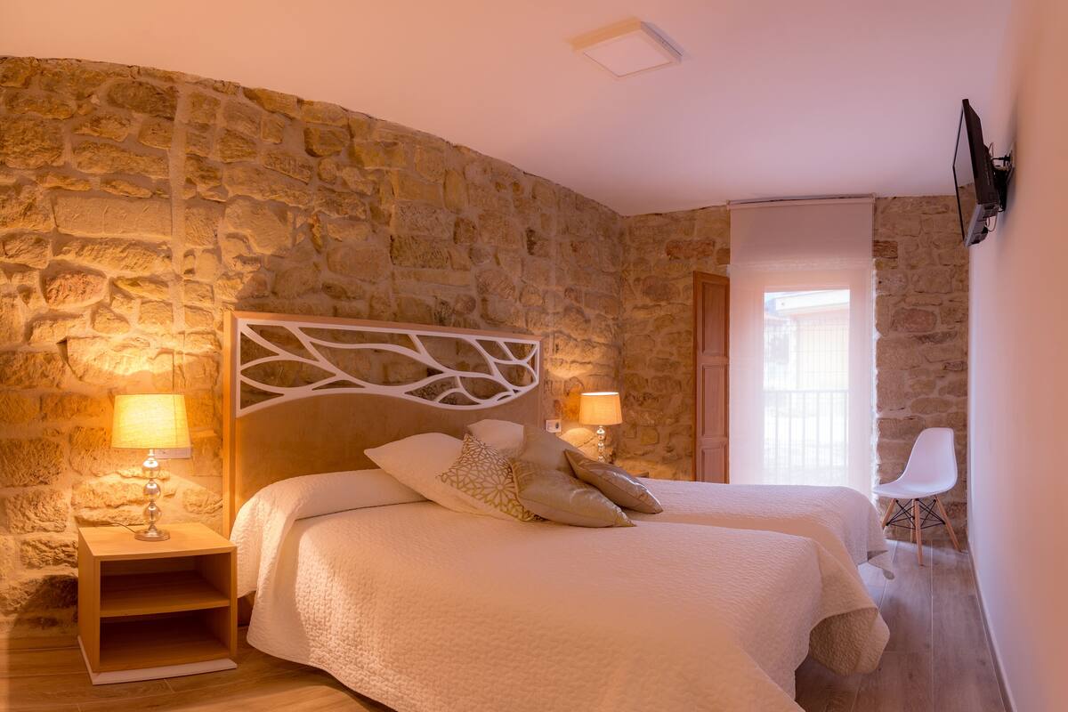 Quarto com parede de pedra, duas camas, abajures, televisão e piso de madeira.
