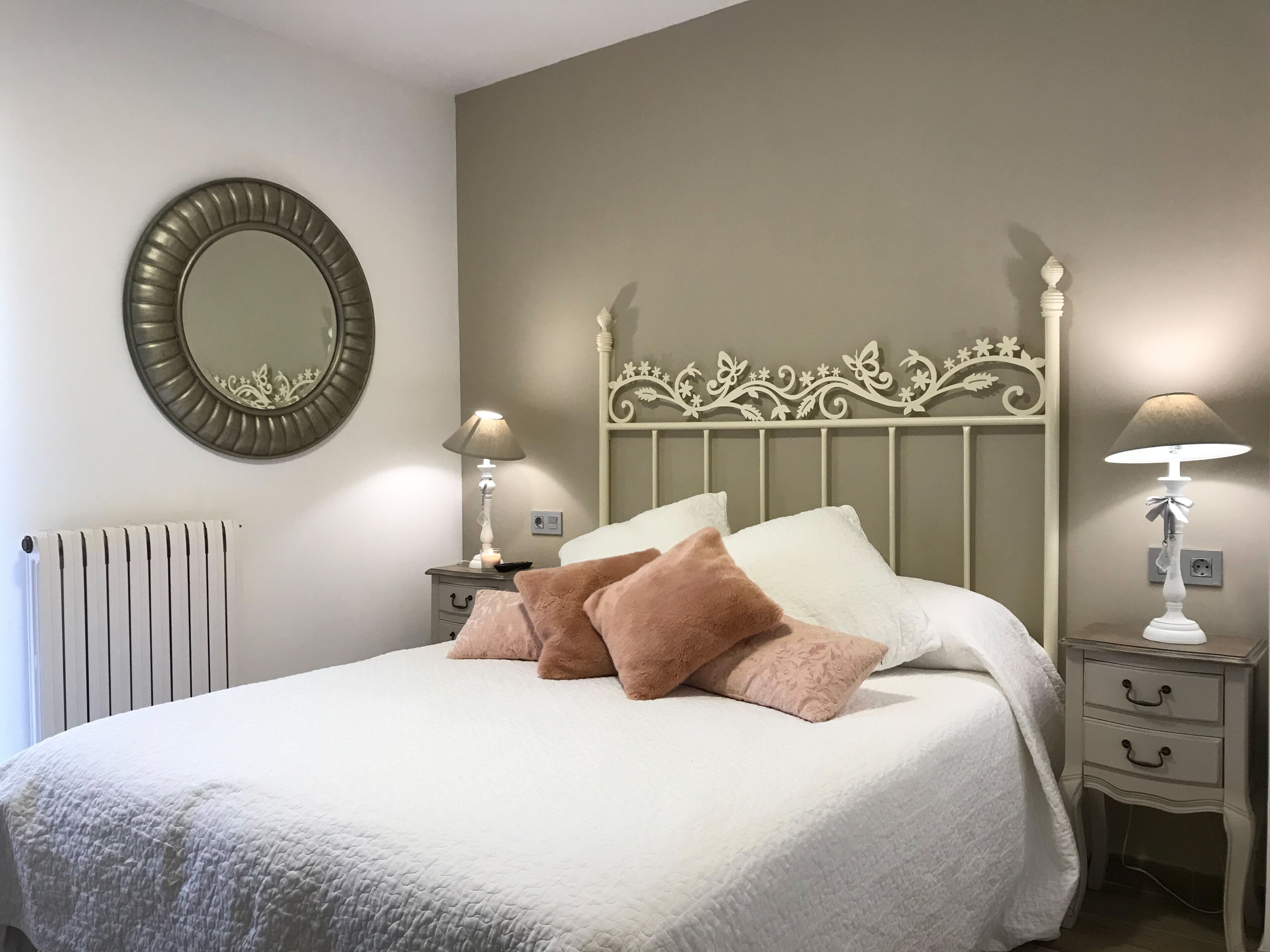 Una cama bien hecha con edredón blanco y varios almohadones, un espejo redondo en la pared y dos mesitas de noche con lámparas.