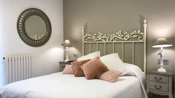 Una cama bien hecha con edredón blanco y varios almohadones, un espejo redondo en la pared y dos mesitas de noche con lámparas.
