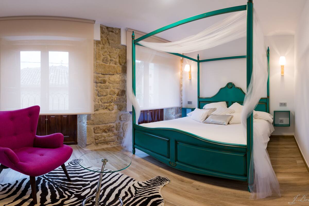 Uma cama de dossel com estrutura azul-turquesa e roupa de cama branca, um tapete com padrão de zebra e uma cadeira rosa vibrante num quarto com paredes de pedra e chão de madeira.