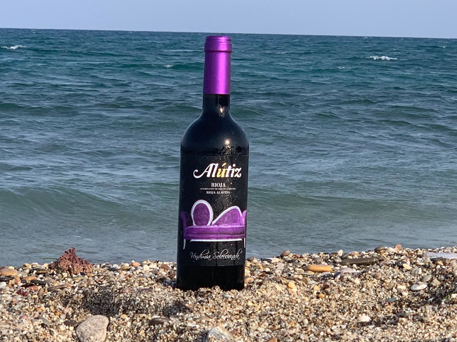 Una botella de vino Alutiz Rioja sobre una playa de guijarros con el océano al fondo.