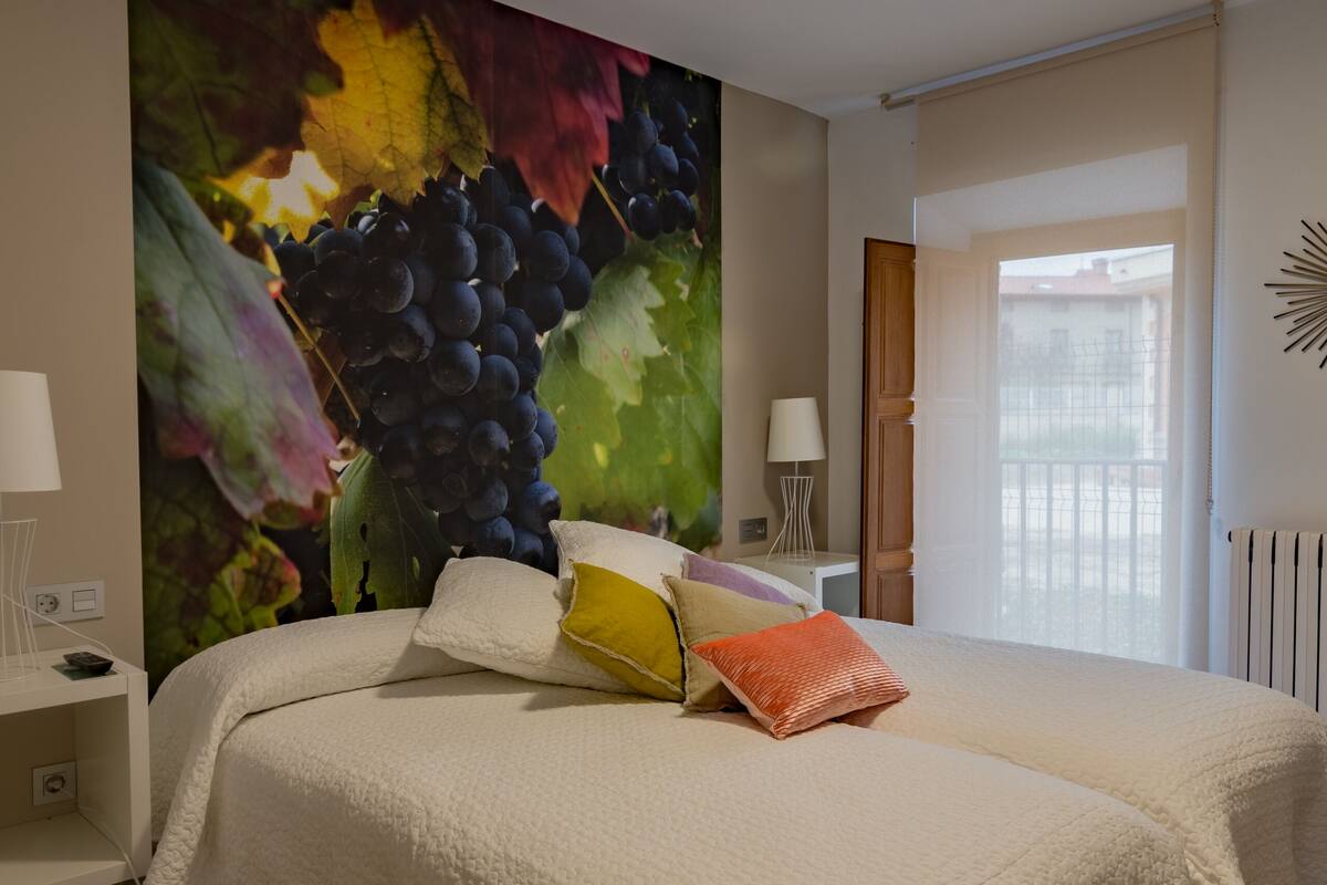 Quarto com uma cama grande, mesa de cabeceira, abajur e um mural na parede com uvas.