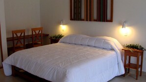 A neatly made bed with white bedding, two wooden chairs, a small table with a plant, and two wall-mounted lamps. - Gocta Andes Lodge (Cocachimba)