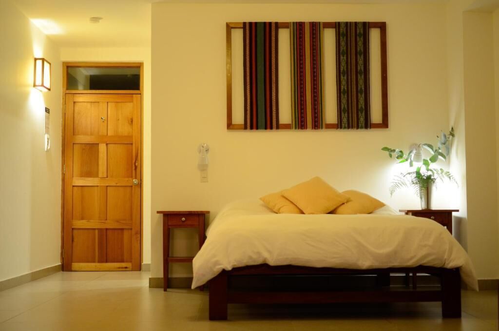 A bedroom with a wooden door, a bed with white bedding, a nightstand, a wall-mounted lamp, and a framed artwork above the bed.