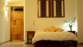 A bedroom with a wooden door, a bed with white bedding, a nightstand, a wall-mounted lamp, and a framed artwork above the bed.