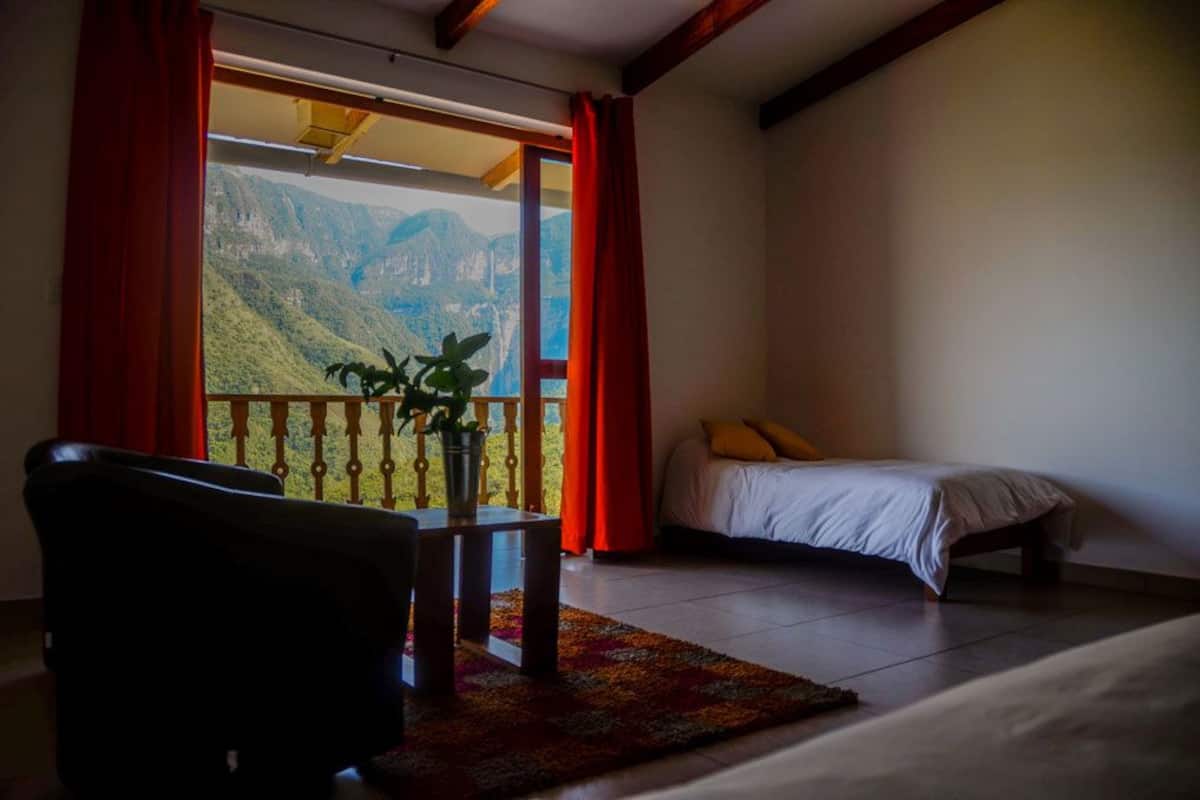 A room with a bed, a table with a plant, and a view of mountains through a large window.