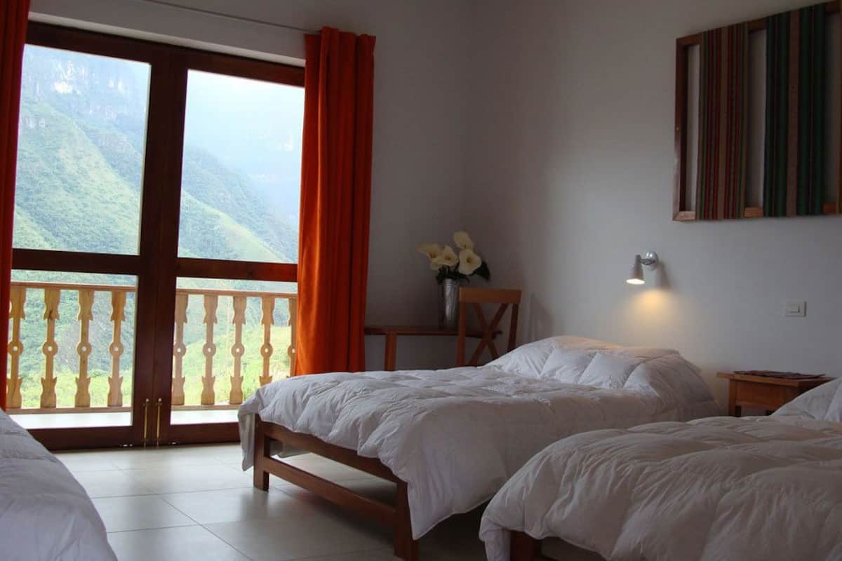 A hotel room with two beds, a balcony with a view of mountains, a vase with flowers, and a wall-mounted light.