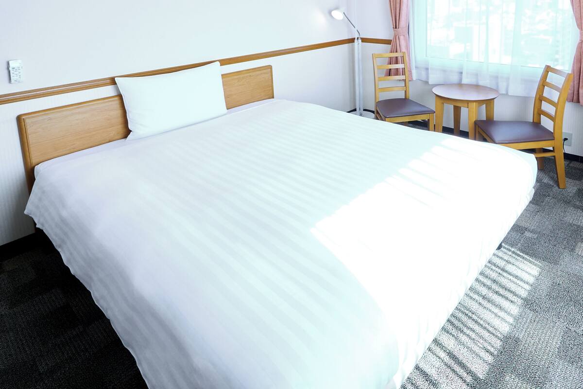 A hotel room with a large bed, a desk with a chair, a lamp, and a window with curtains.