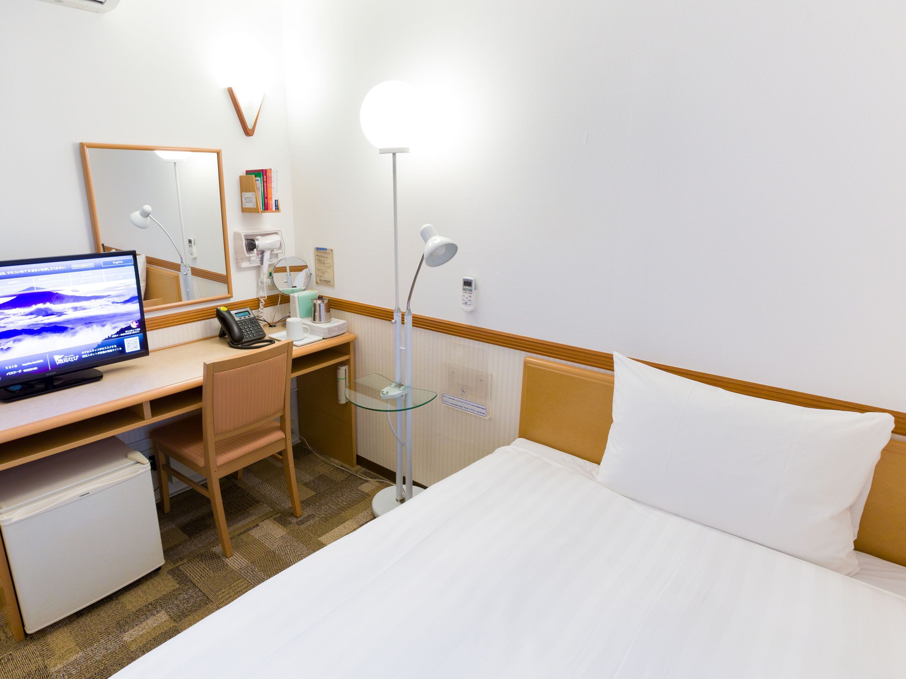 A hotel room with a bed, a desk with a computer, a chair, a small refrigerator, a bedside table, a lamp, and a mirror.