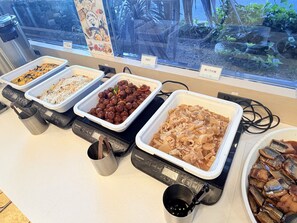A buffet with various dishes in white trays, a Panasonic rice cooker, and a view of greenery through a window.