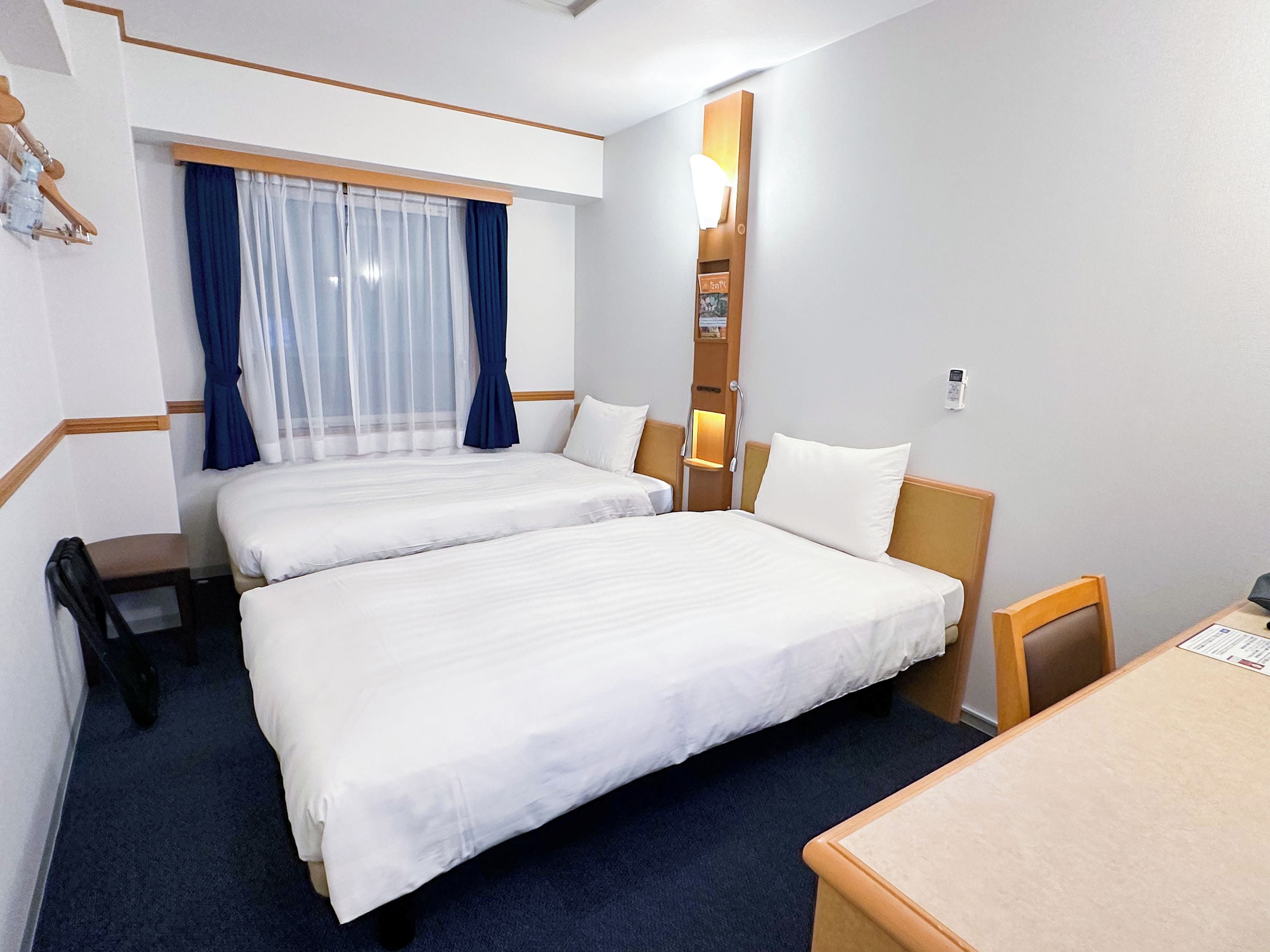 A hotel room with two beds, a desk, a chair, a nightstand, a lamp, and a window with curtains.