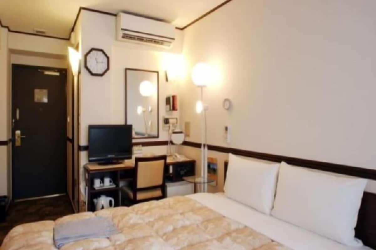 A hotel room with a bed, a desk with a television, a mirror, and a wall-mounted air conditioner.