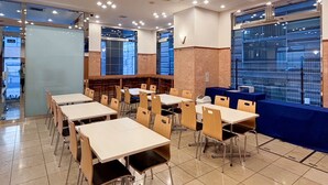 Een ruime eetruimte met meerdere tafels en stoelen, grote ramen met blauw getinte glas en een modern plafond met inbouwverlichting.
