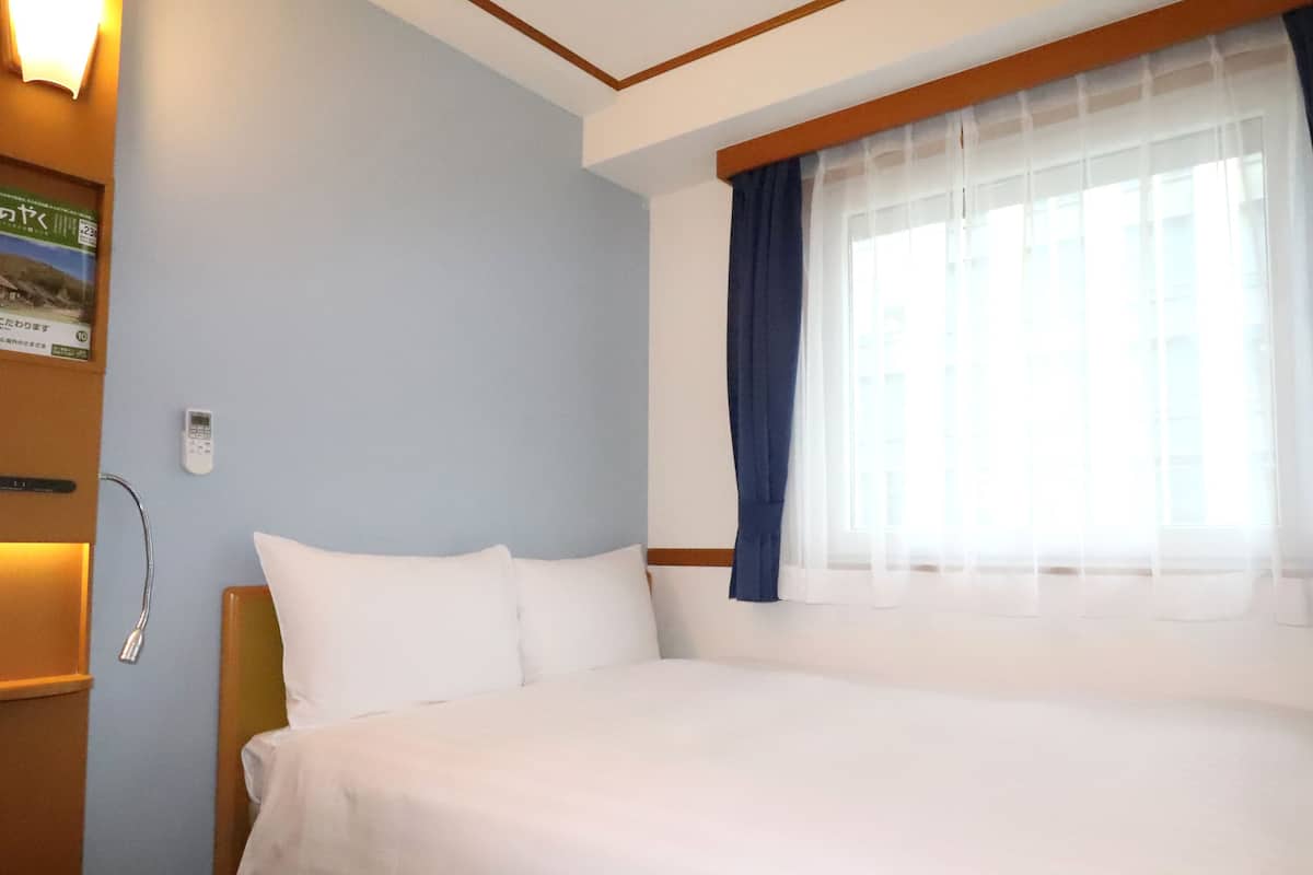 Ein Hotelzimmer mit Bett, Nachttisch, Lampe, einem Fenster mit Vorhängen und einem an der Wand montierten Telefon.