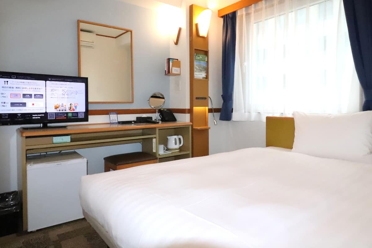 Ein Hotelzimmer mit einem Bett, einem Schreibtisch mit Computer, einem kleinen Kühlschrank, einer Kaffeemaschine und einem Fenster mit Vorhängen.