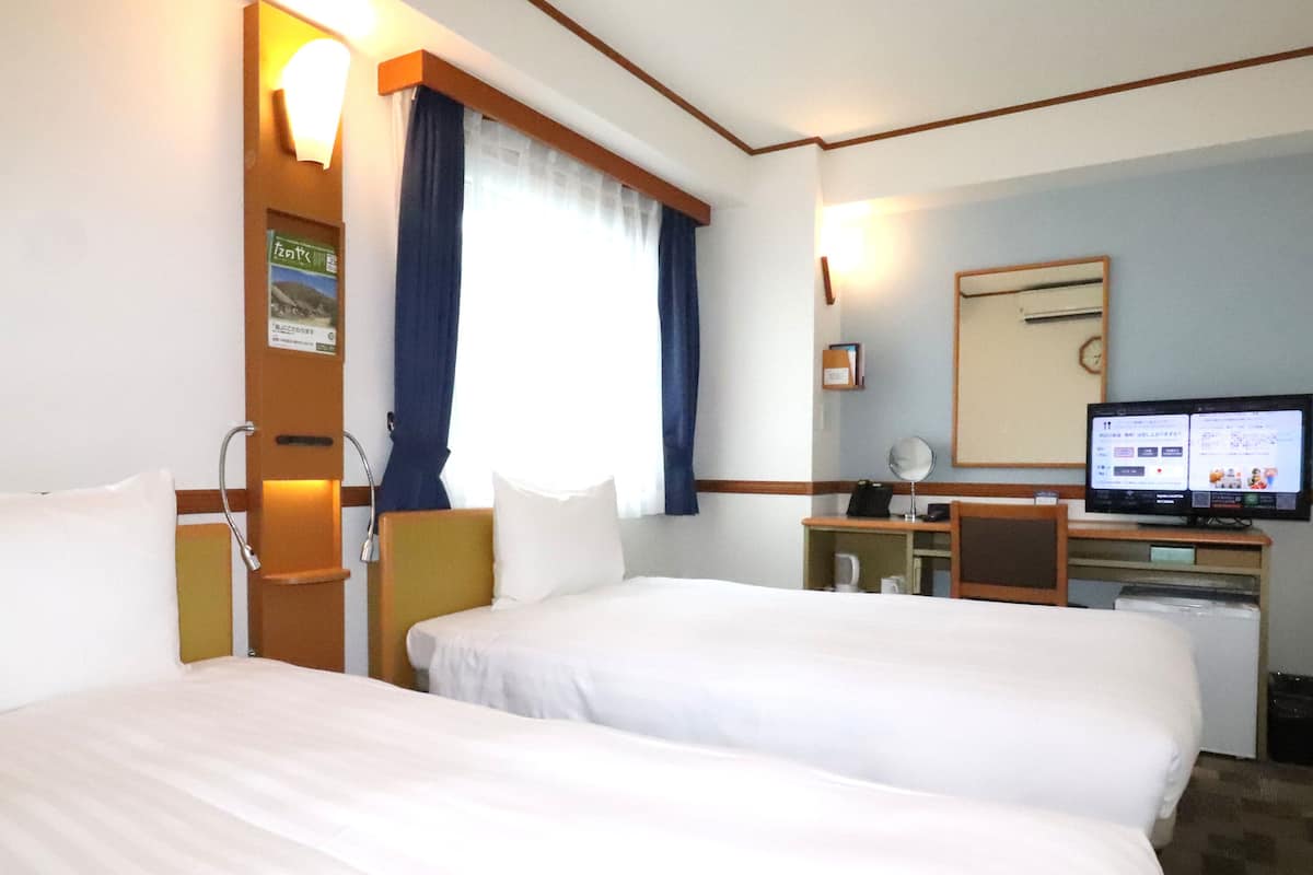 Ein Hotelzimmer mit zwei Betten, einem Schreibtisch mit Computer, einem Spiegel und einem Fenster mit Vorhängen.