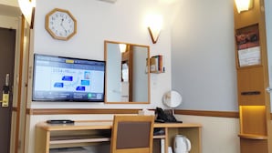 Quarto de hotel com escrivaninha, cadeira, cama, televisão, relógio, espelho e uma pequena estante com livros e chaleira.