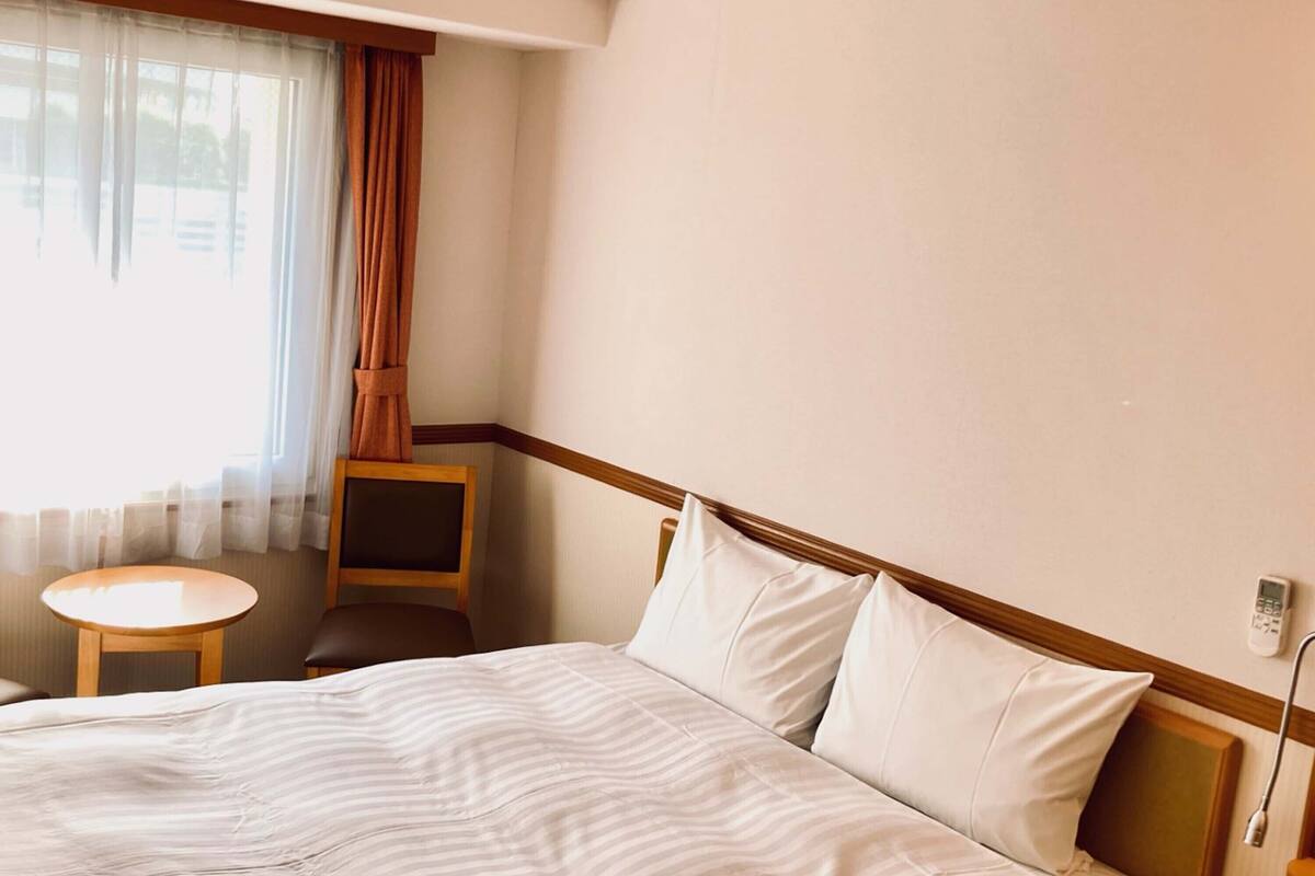 A hotel room with a bed, a small round table, a chair, and a window with curtains.